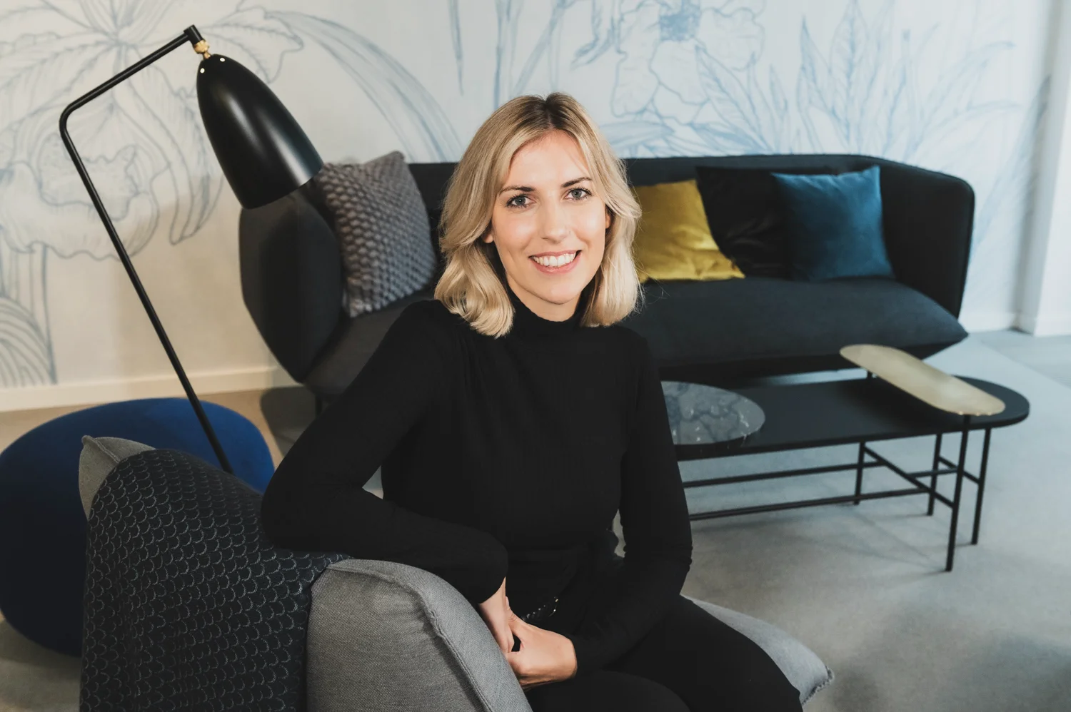 Smiling employee in a black turtleneck sitting in the lounge with botanical wallpaper, business portrait, Frankfurt am Main & Munich — Digital+, corporate photography by Janina Laszlo
