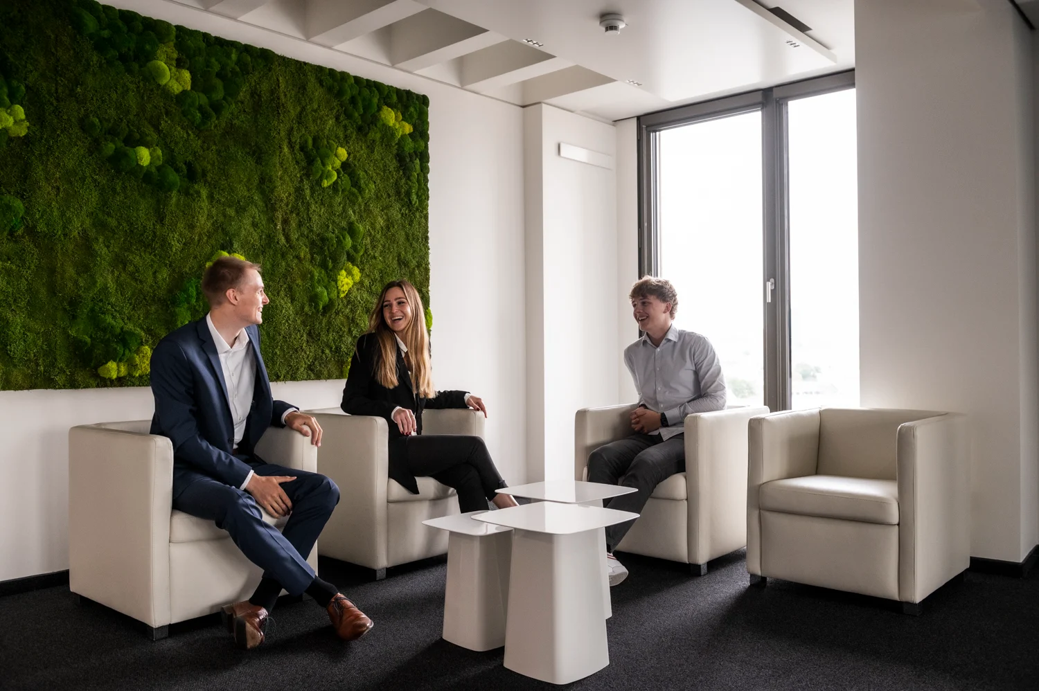 Three colleagues laughing in white leather armchairs in front of a moss wall, Frankfurt am Main — Digital+, corporate photography by Janina Laszlo
