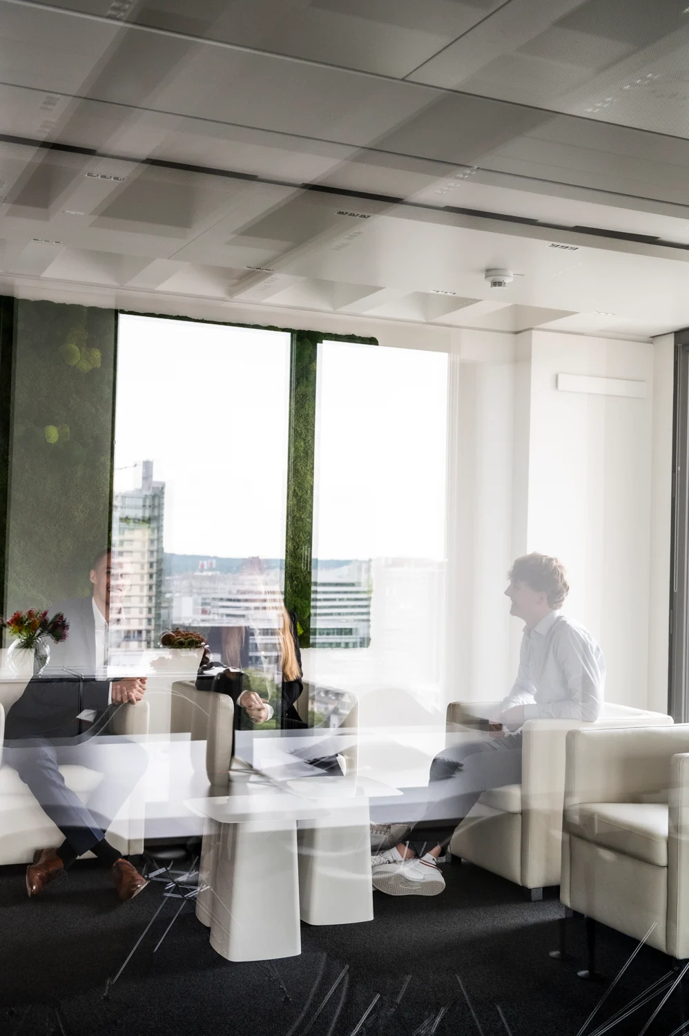 Three colleagues in the moss-wall lounge seen through reflecting glass, high-rise view behind, Frankfurt am Main — Digital+, corporate photography by Janina Laszlo