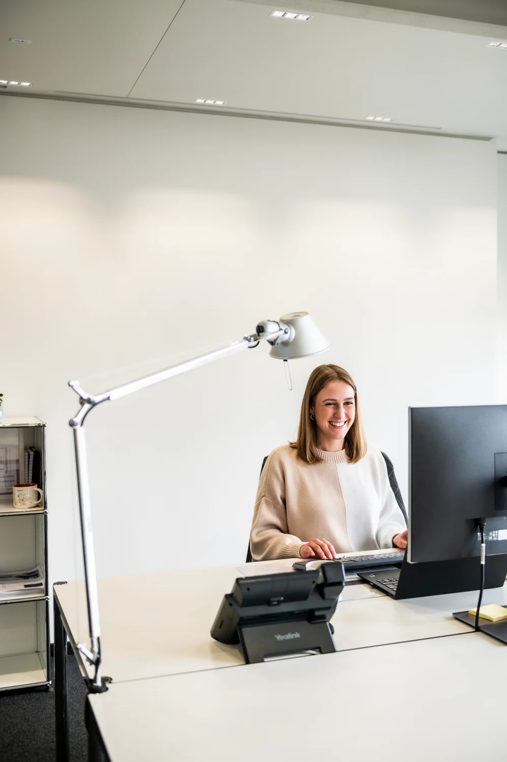 Smiling employee at her desk with a Tolomeo lamp and monitor, bright white office, Frankfurt am Main — Digital+, corporate photography by Janina Laszlo