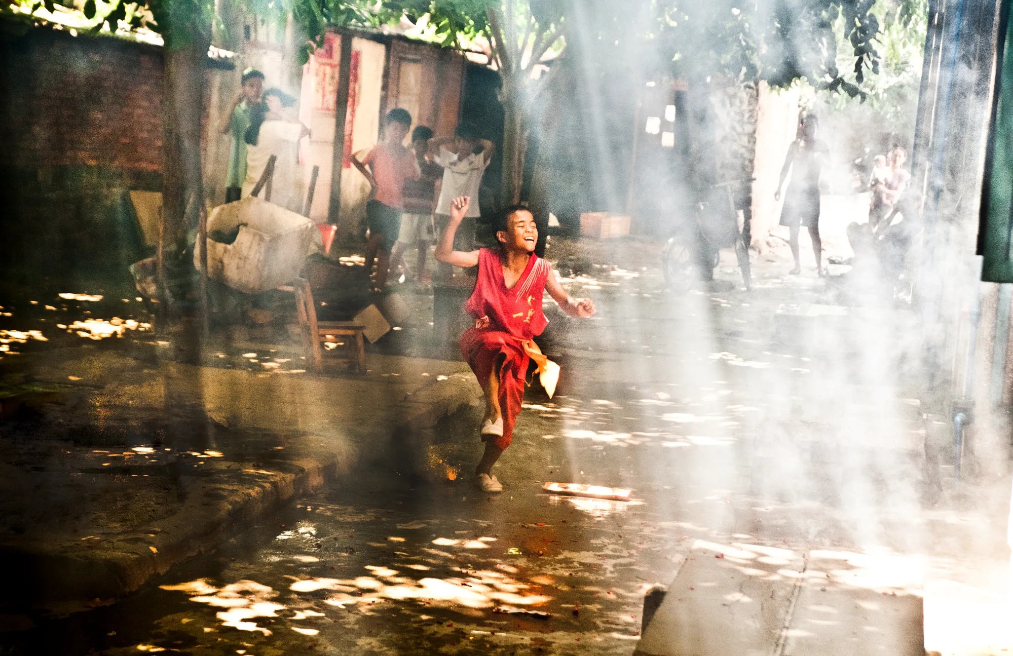 Boy in red running through sunbeams and smoke in a village street — Dragon Boat Festival Hainan, documentary photography by Janina Laszlo