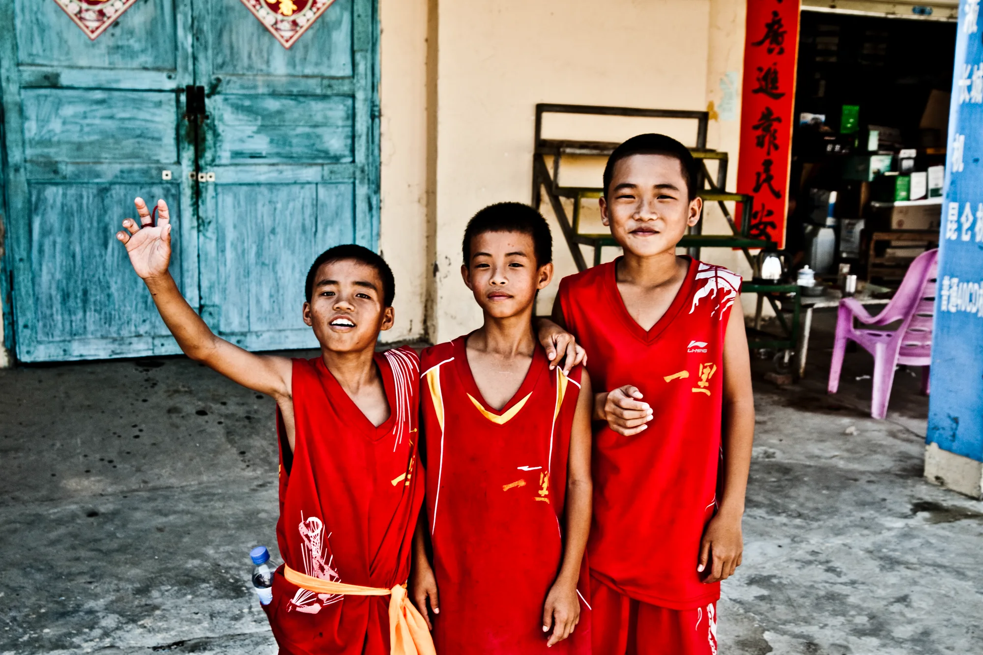 Three boys in red festival shirts waving in front of a blue door — Dragon Boat Festival Hainan, documentary photography by Janina Laszlo
