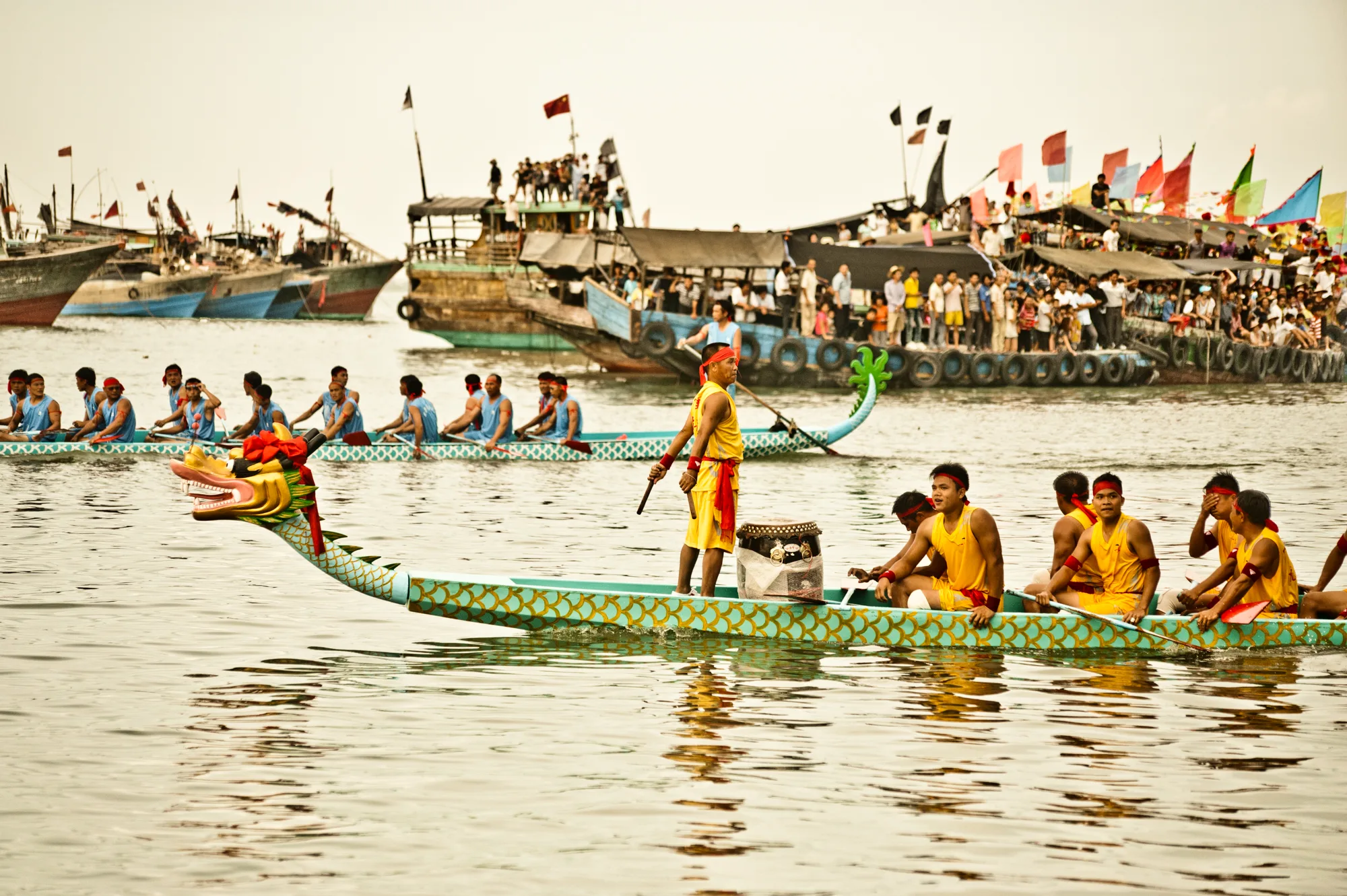 Dragon boat crew in yellow rowing past boats decked with flags — Dragon Boat Festival Hainan, documentary photography by Janina Laszlo