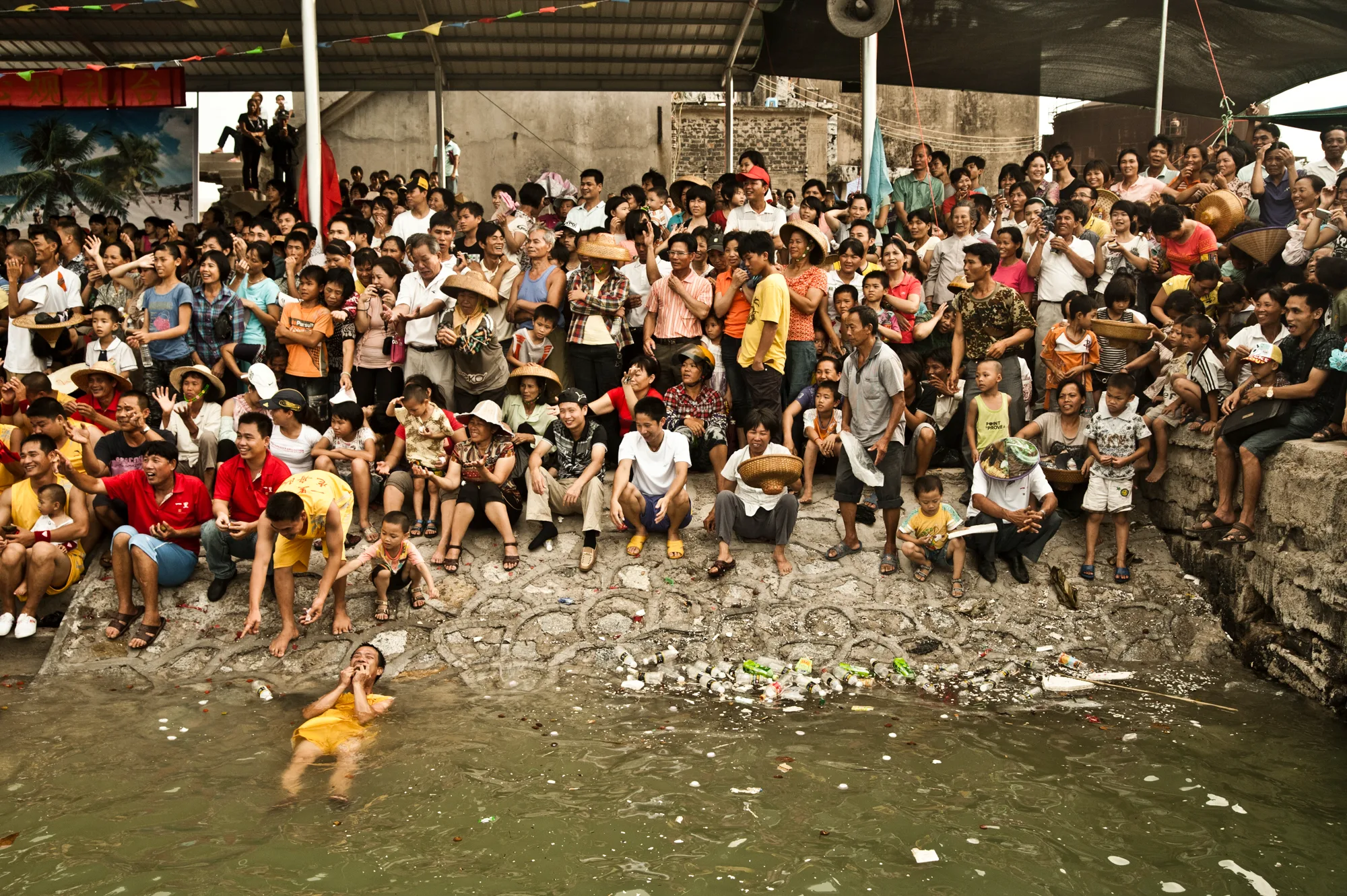 Dense crowd of spectators on the shore, a swimmer in the water — Dragon Boat Festival Hainan, documentary photography by Janina Laszlo