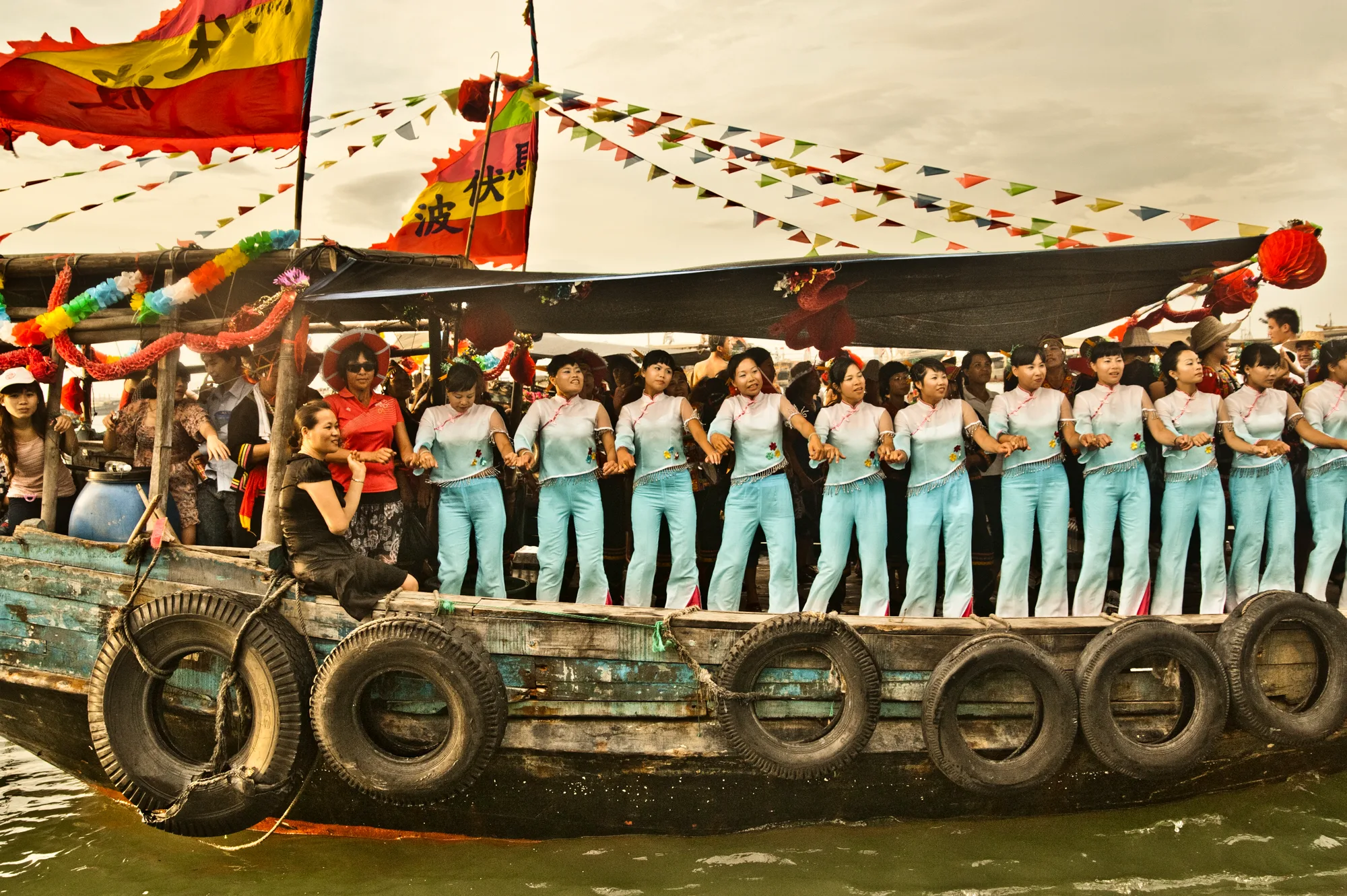 Decorated boat with tyre fenders carrying dancers in blue — Dragon Boat Festival Hainan, documentary photography by Janina Laszlo