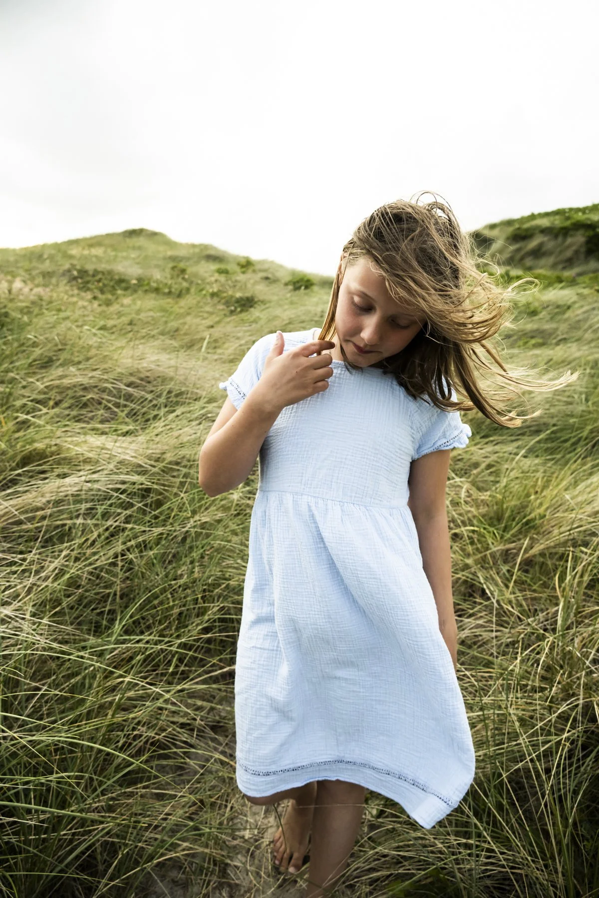 Girl in a light blue summer dress standing in windy dune grass, Klitmøller, Ernsting's family kidswear — campaign photography by Janina Laszlo, Munich