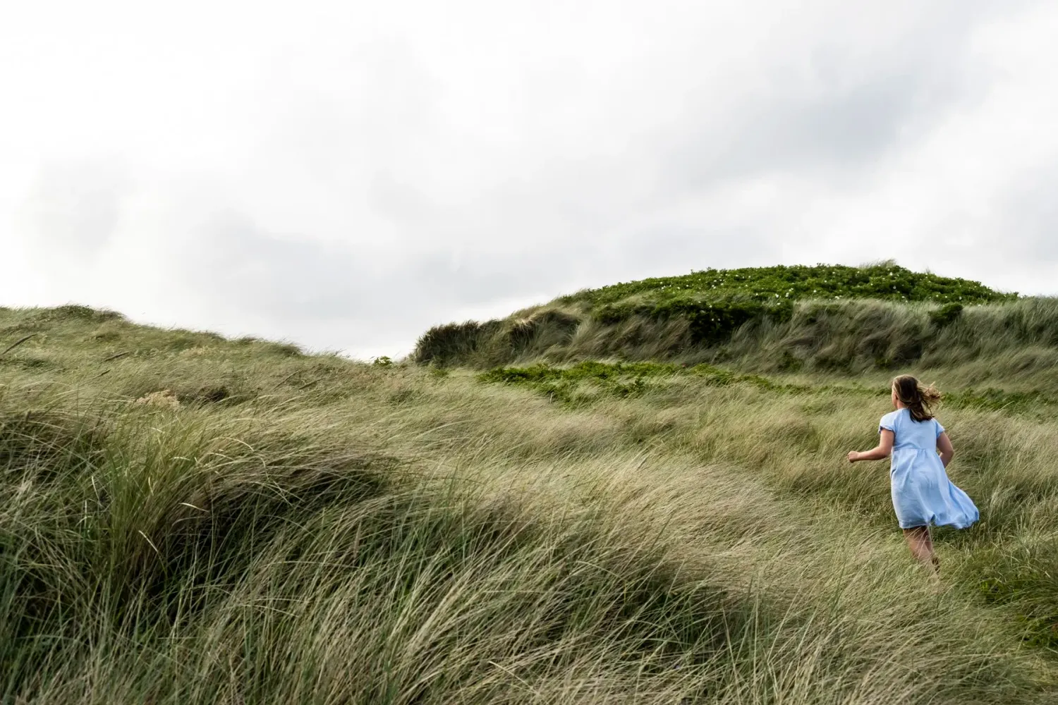 Child running through tall dune grass under a cloudy sky — Ernsting's family kidswear, Klitmøller, Denmark — campaign photography by Janina Laszlo, Munich
