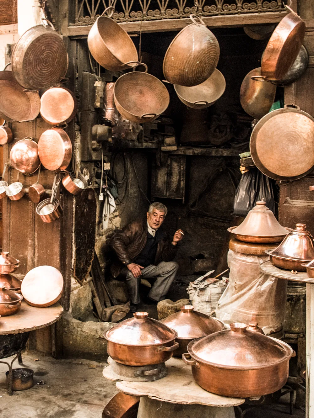 Coppersmith resting in his workshop among hammered copper pots and tagines, Fez medina — Fez, street and travel photography by Janina Laszlo