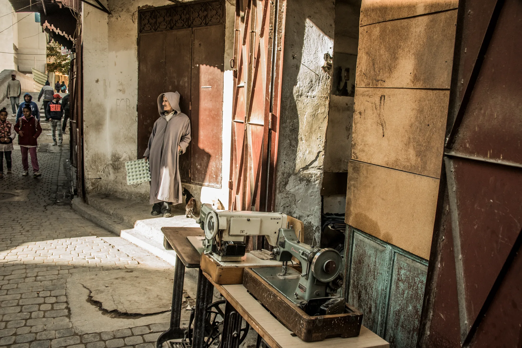 Old man in a hooded djellaba beside vintage sewing machines in a medina lane, Fez — Fez, street and travel photography by Janina Laszlo