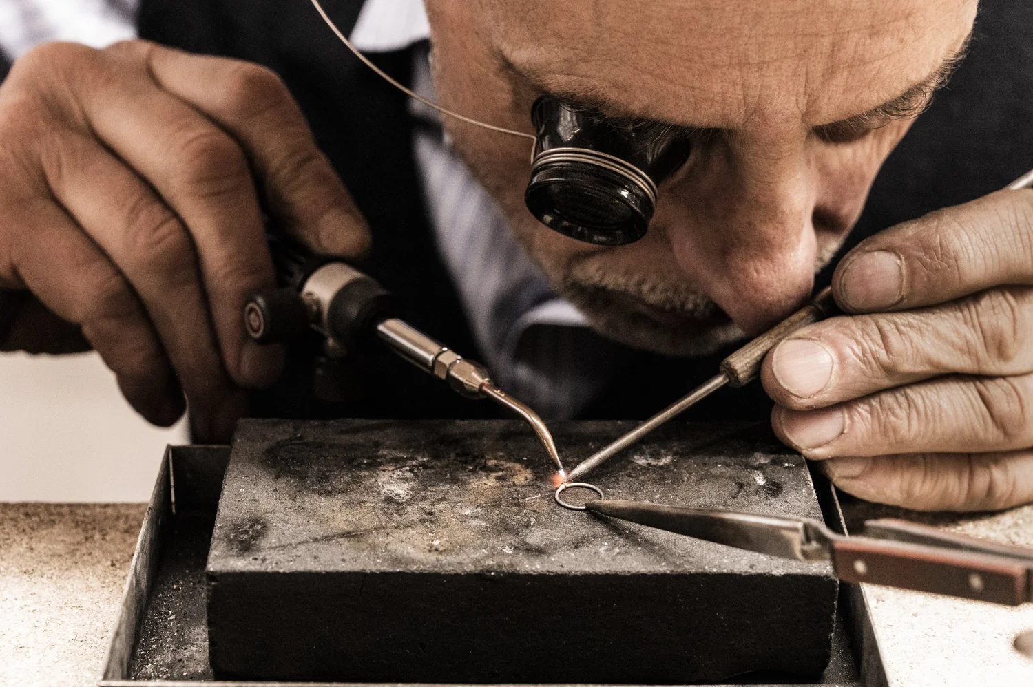 Goldsmith Gerd Weickmann soldering a piece of jewellery with a fine flame, loupe on his eye — reportage by Janina Laszlo, photographer in Munich