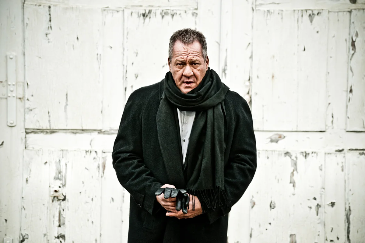 Hugo Bachmaier in a dark coat holding a camera against a white wall — Hugo Bachmaier, portrait photography by Janina Laszlo