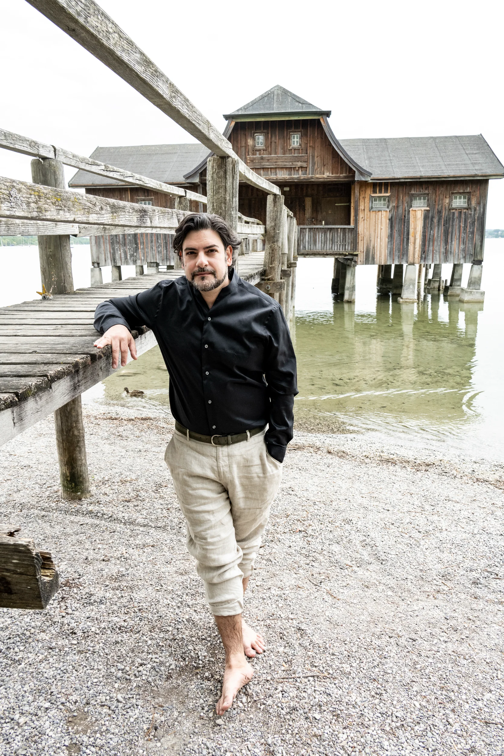 Ilker Arcayürek leaning on a wooden jetty in front of a boathouse — Ilker Arcayürek, artist portrait photography by Janina Laszlo