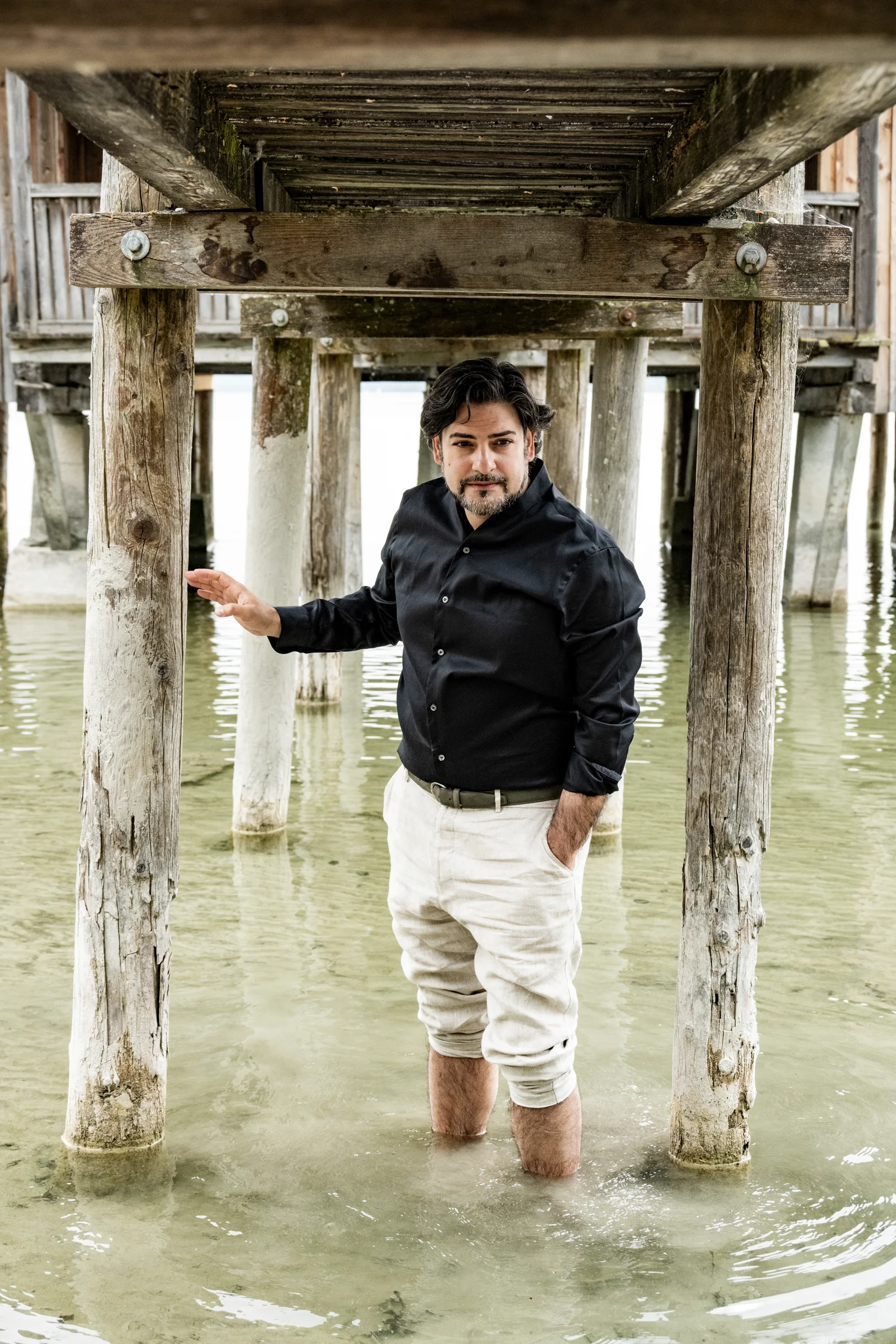 Ilker Arcayürek standing in the water under a wooden jetty — Ilker Arcayürek, artist portrait photography by Janina Laszlo