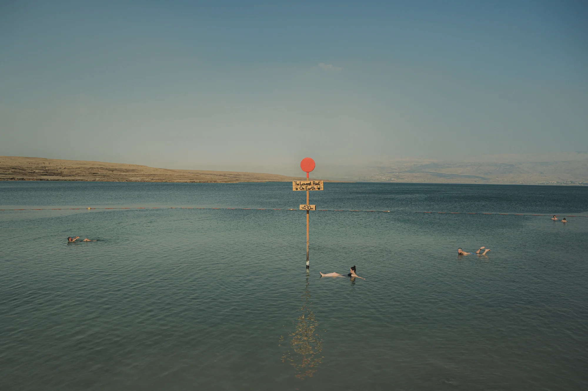 Bathers floating in the still turquoise water around a red-topped pole marked "The Lowest Point on Earth −430 m", Dead Sea, Israel — Israel, travel and street photography by Janina Laszlo