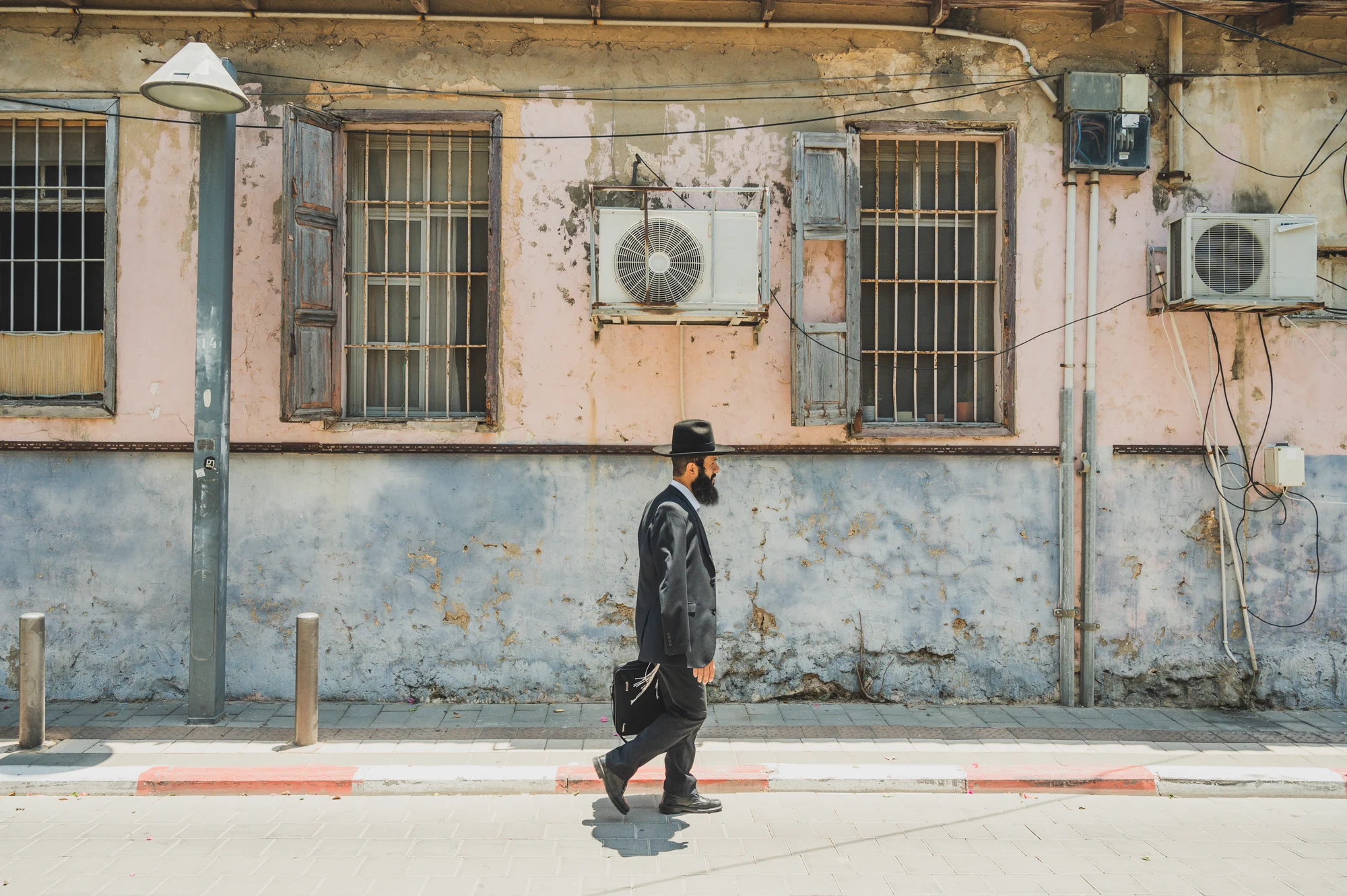 Orthodox Jewish man in black hat and suit walking past a weathered pink and blue facade with barred windows and air conditioners, Tel Aviv — Israel, travel and street photography by Janina Laszlo