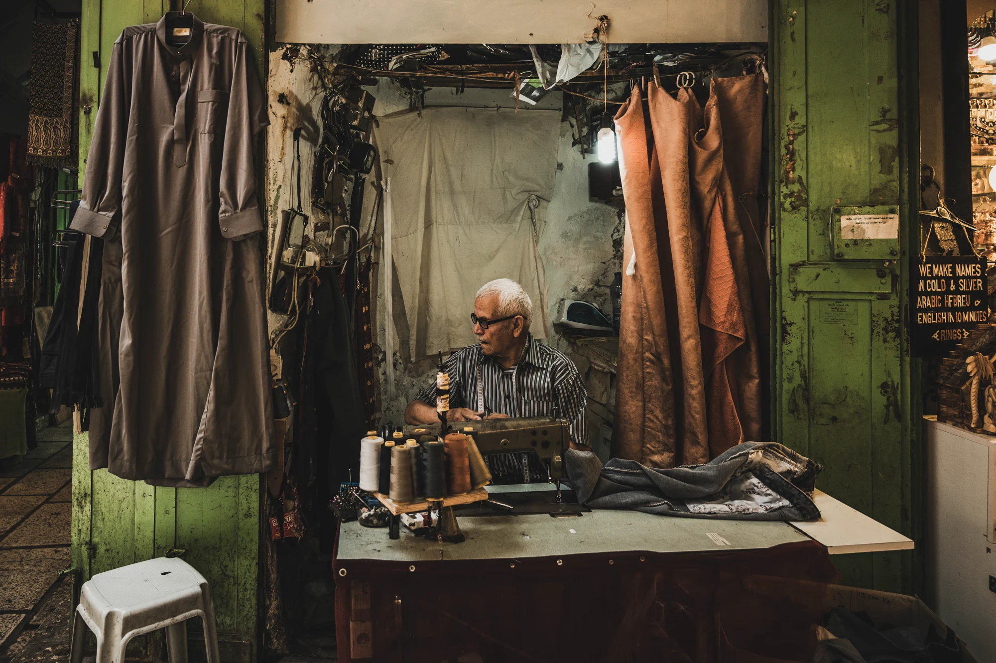 Elderly tailor at his sewing machine in a green wooden market stall hung with robes, Old City of Jerusalem — Israel, travel and street photography by Janina Laszlo