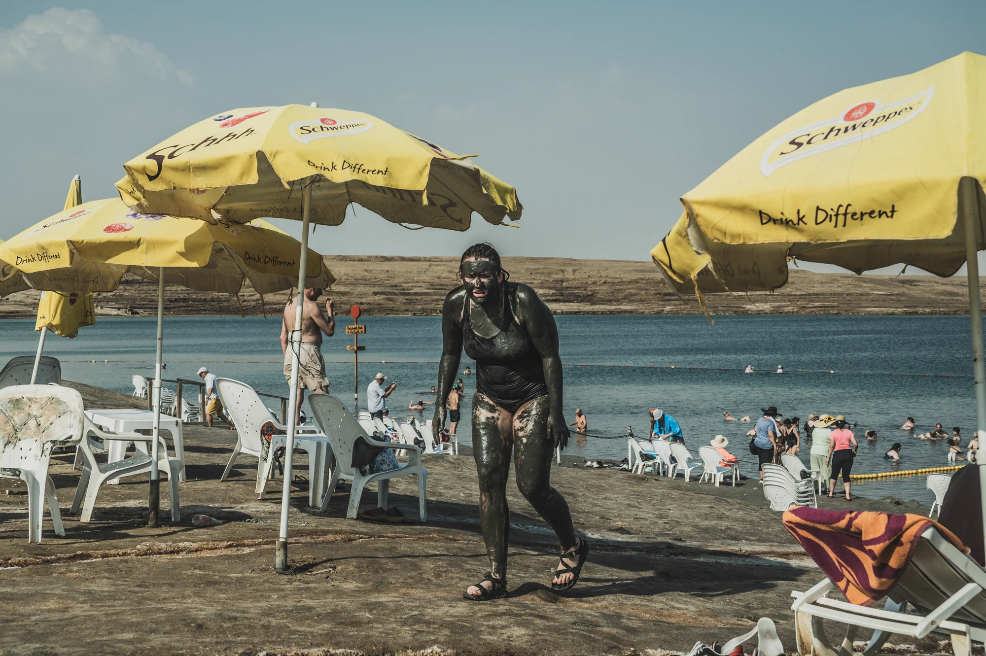 Woman covered head to toe in black mud walking past yellow beach umbrellas, bathers in the water behind, Dead Sea, Israel — Israel, travel and street photography by Janina Laszlo