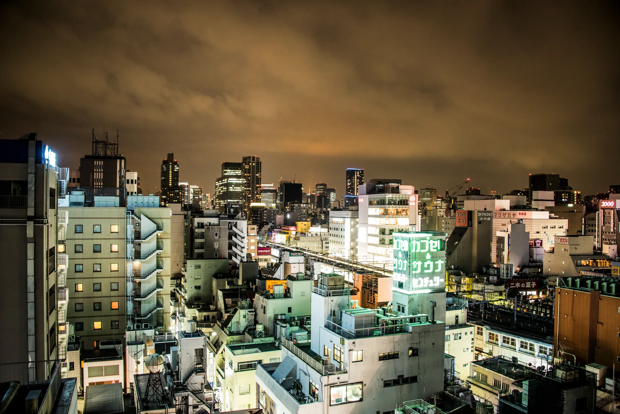 Night view over lit office blocks and a neon capsule hotel sign, Tokyo — Japan, travel and street photography by Janina Laszlo