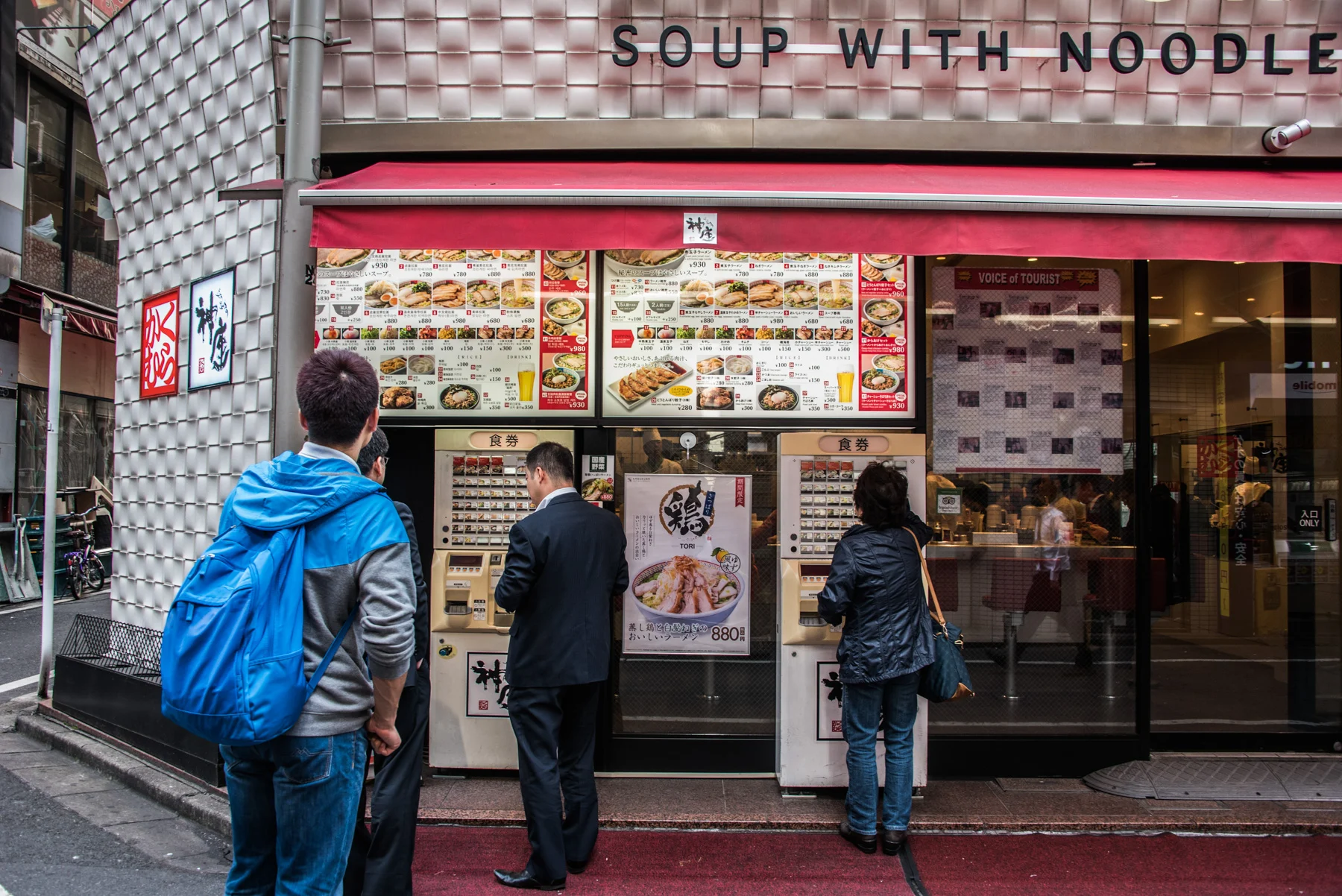 Customers buying meal tickets at vending machines outside a ramen shop under a red awning, Tokyo — Japan, travel and street photography by Janina Laszlo