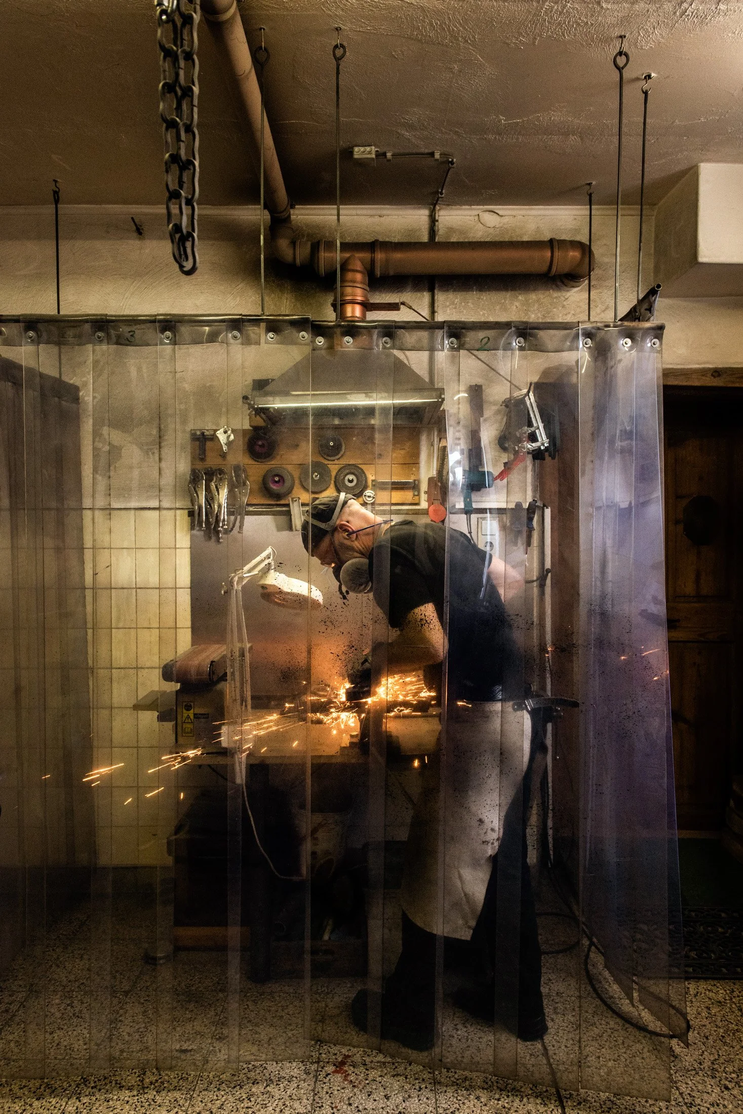 Knife smith seen through a glass partition while grinding a blade — reportage by Janina Laszlo, photographer in Munich
