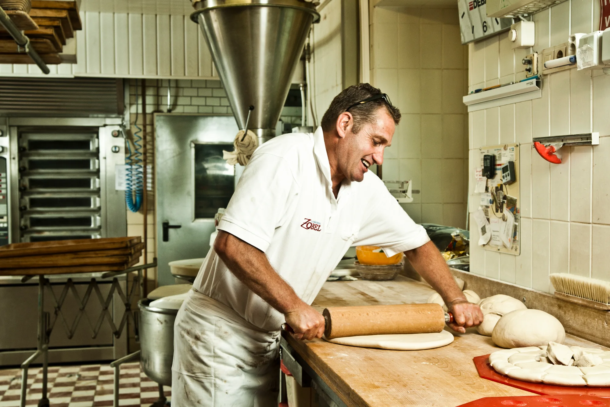 Baker in a Zobel polo shirt rolling out dough with a wooden rolling pin at the workbench — Landbäckerei Zobel, Seefeld, Bavaria, craft bakery photography by Janina Laszlo