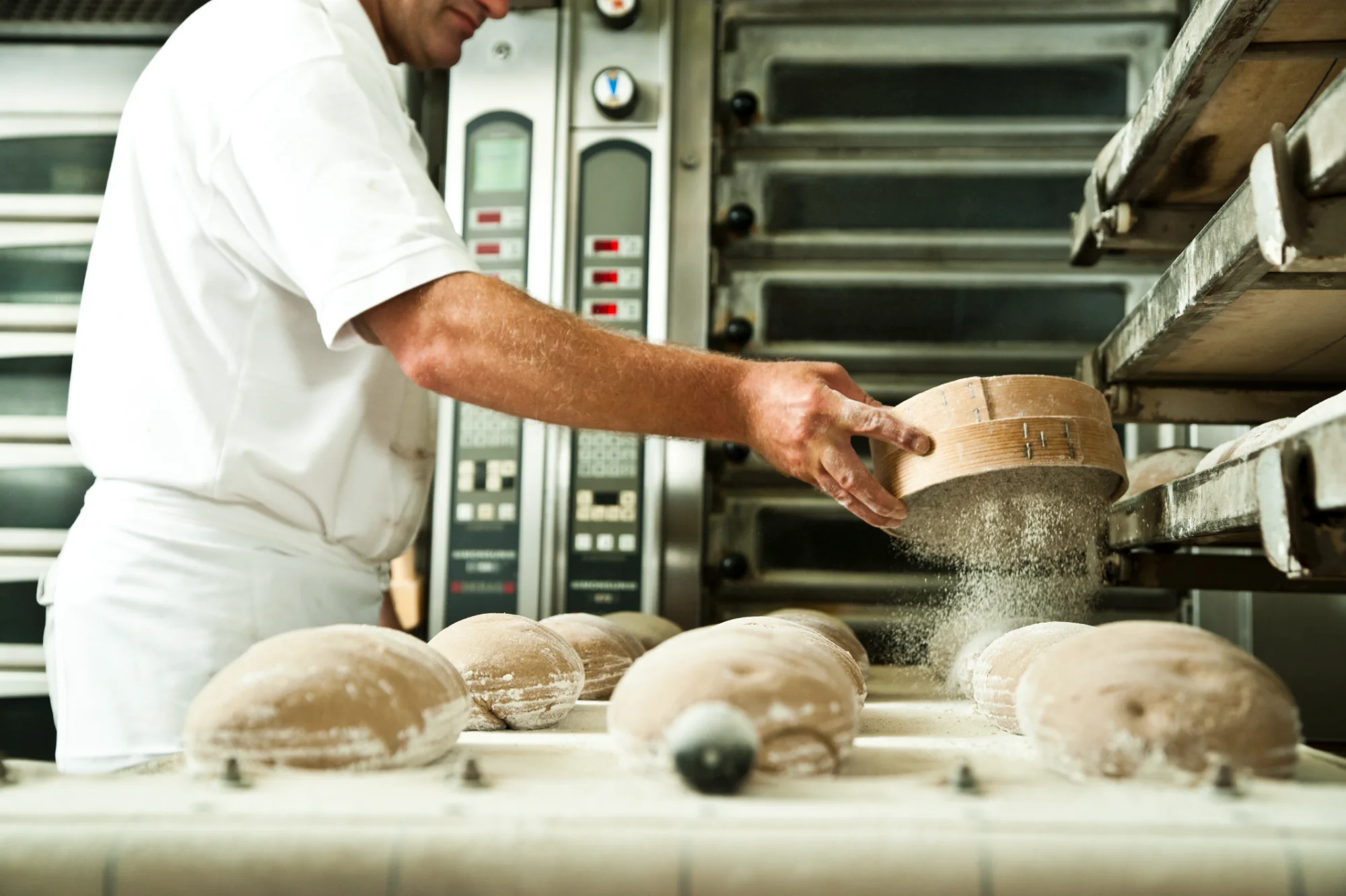 Baker dusting flour through a wooden sieve over proofing loaves in front of the deck ovens — Landbäckerei Zobel, Seefeld, Bavaria, craft bakery photography by Janina Laszlo