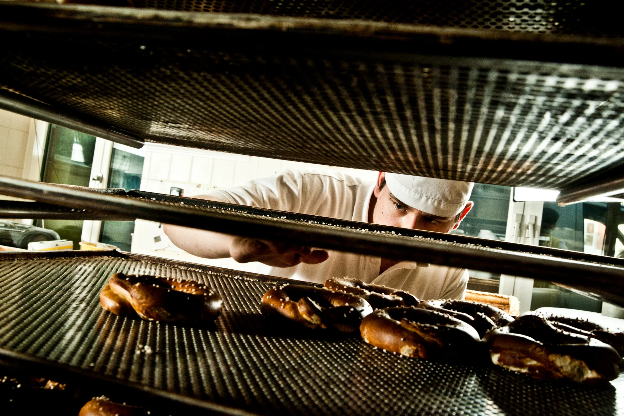 Young baker in a white cap reaching for salted pretzels on perforated baking trays, view through the rack — Landbäckerei Zobel, Seefeld, Bavaria, craft bakery photography by Janina Laszlo