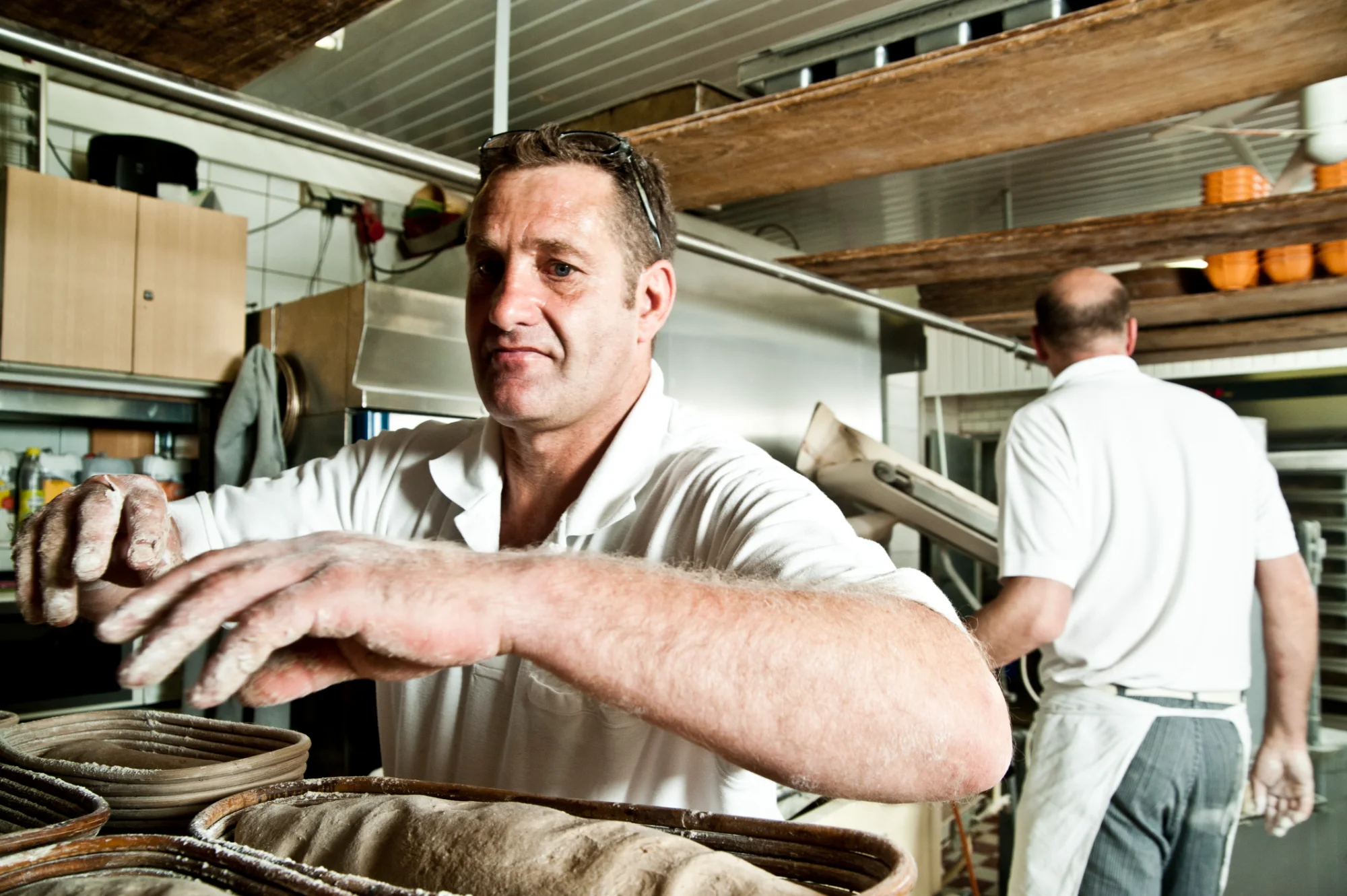 Baker with flour-covered hands at the proofing baskets, colleague working in the background — Landbäckerei Zobel, Seefeld, Bavaria, craft bakery photography by Janina Laszlo
