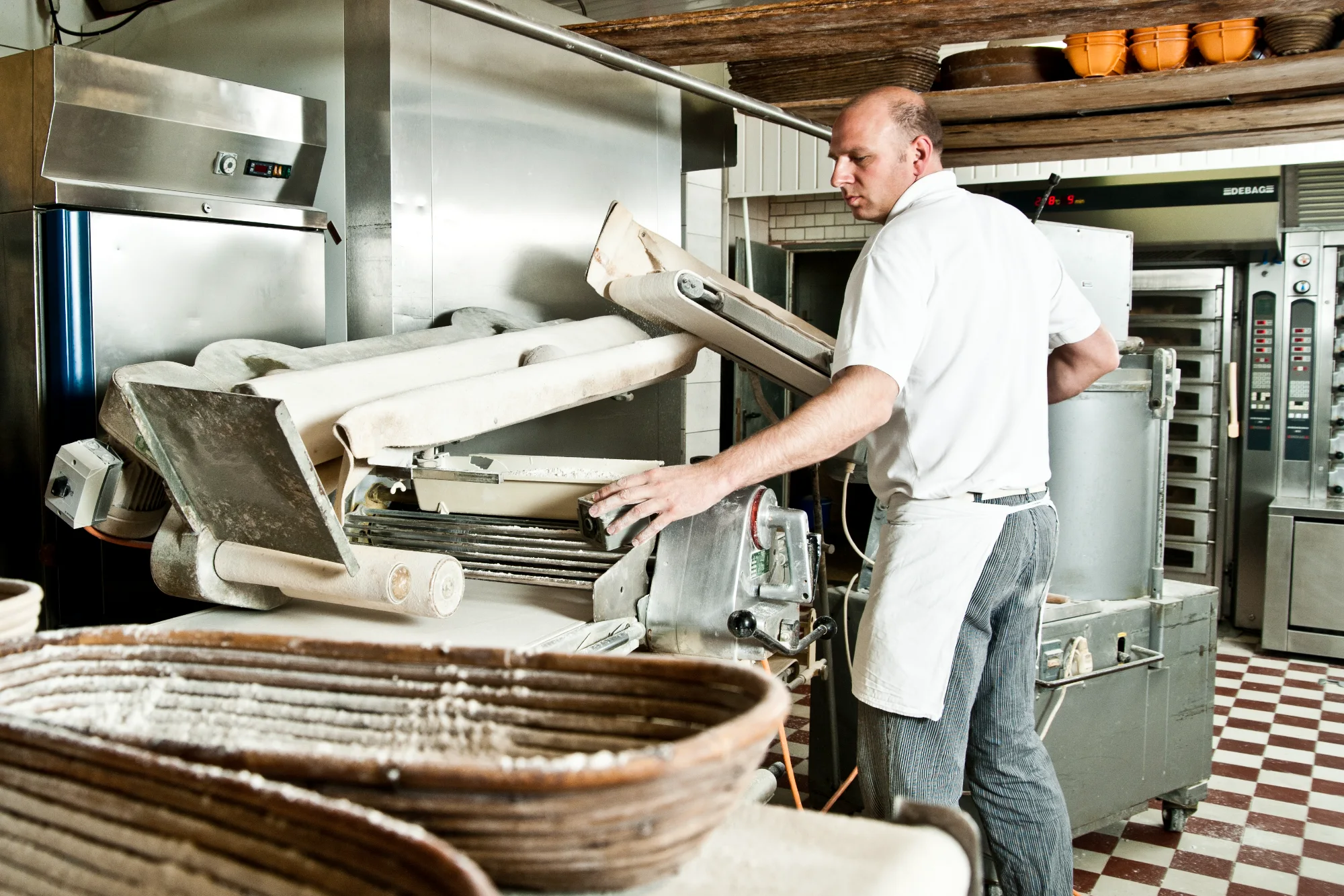 Baker feeding dough into a dough sheeter, floured proofing basket in the foreground — Landbäckerei Zobel, Seefeld, Bavaria, craft bakery photography by Janina Laszlo