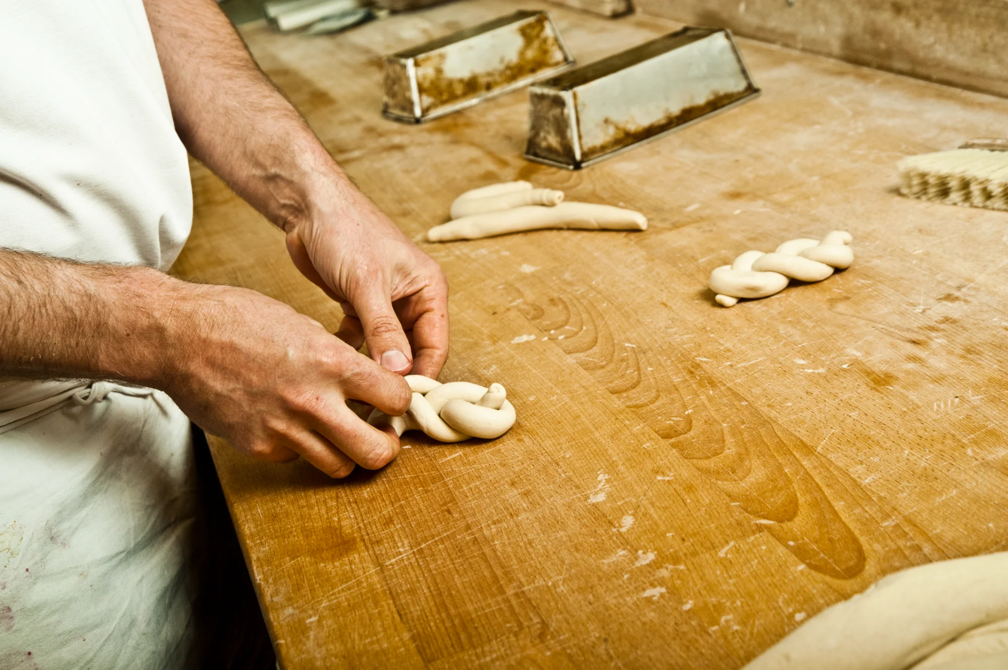 Hands braiding a small plaited dough roll on a worn wooden workbench — Landbäckerei Zobel, Seefeld, Bavaria, craft bakery photography by Janina Laszlo