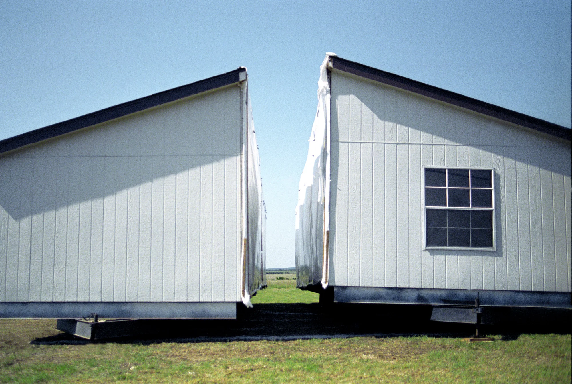 Two halves of a white mobile home standing apart on a lawn, a strip of prairie visible between them — Love County, Oklahoma, analog documentary photography by Janina Laszlo