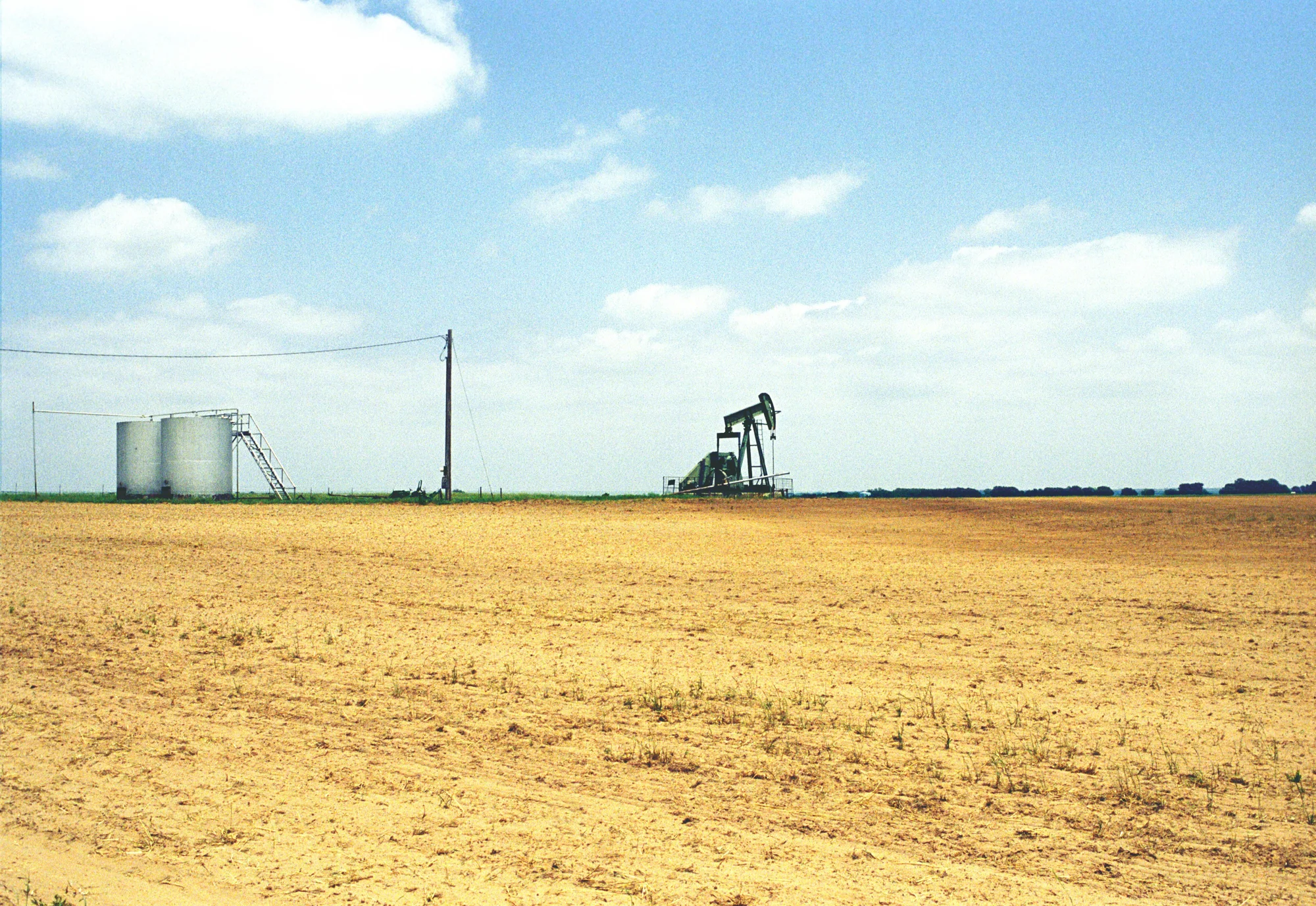 Oil pumpjack and storage tanks on a bare, sun-baked field under a wide sky — Love County, Oklahoma, analog documentary photography by Janina Laszlo