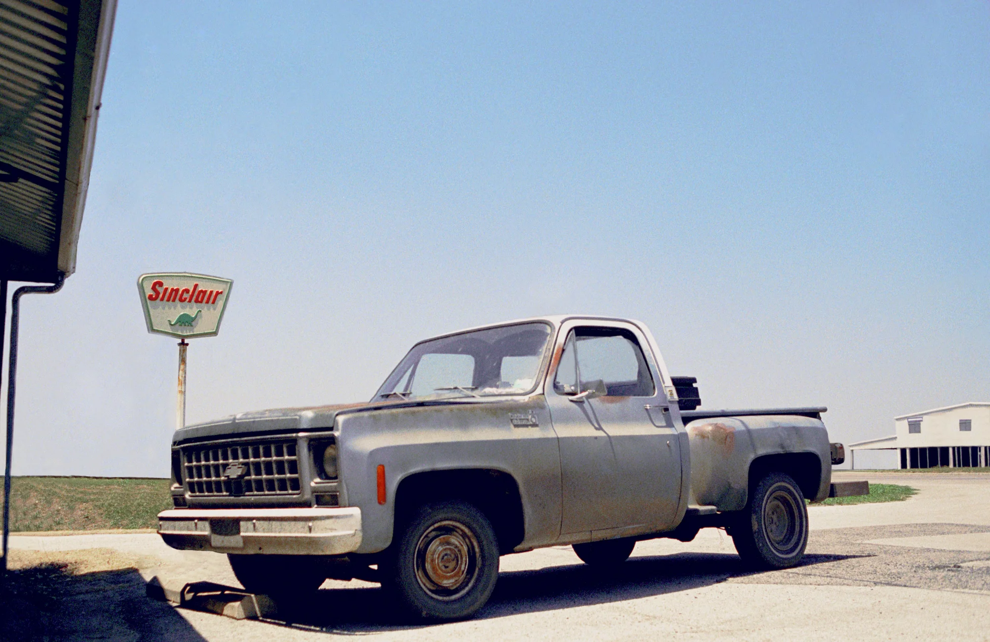 Rusty silver Chevrolet pickup parked beside a vintage Sinclair dinosaur sign — Love County, Oklahoma, analog documentary photography by Janina Laszlo