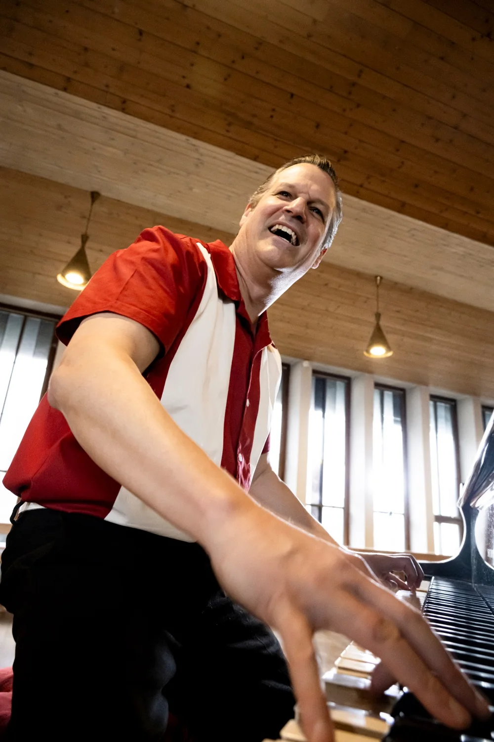 Matthias Heiligensetzer laughing at the piano in a red and white shirt, low angle — Matthias Heiligensetzer, pianist & entertainer, musician portrait photography by Janina Laszlo, Munich