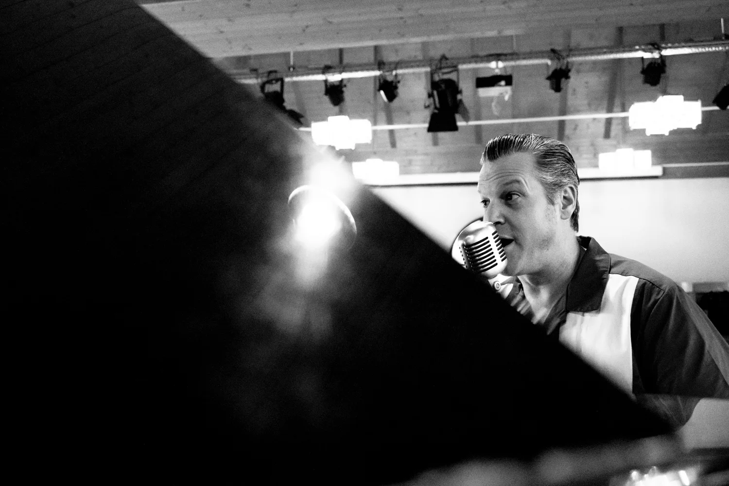 Matthias Heiligensetzer singing behind the raised lid of a grand piano under stage lights, black-and-white — Matthias Heiligensetzer, pianist & entertainer, musician portrait photography by Janina Laszlo, Munich