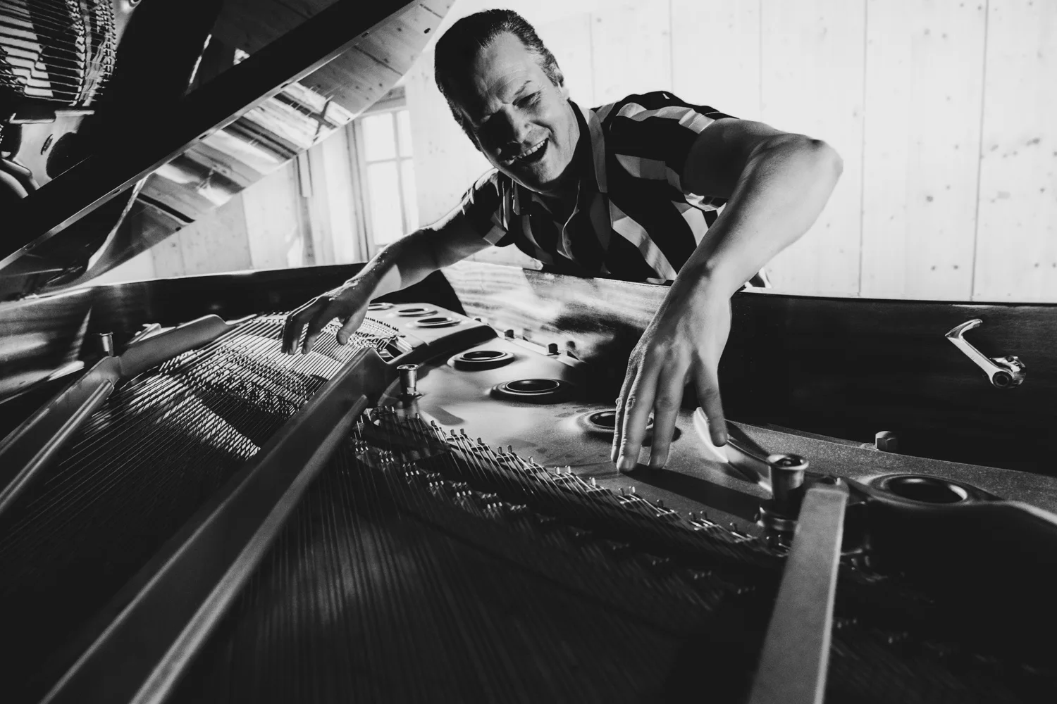 Matthias Heiligensetzer laughing as he reaches into the strings of a grand piano, black-and-white — Matthias Heiligensetzer, pianist & entertainer, musician portrait photography by Janina Laszlo, Munich