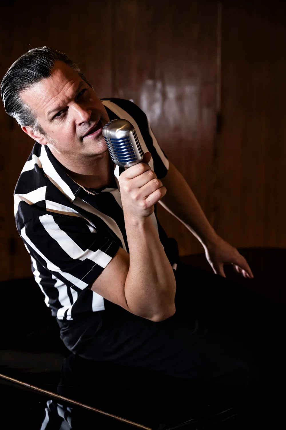Matthias Heiligensetzer in a striped shirt singing into a vintage microphone against dark wood — Matthias Heiligensetzer, pianist & entertainer, musician portrait photography by Janina Laszlo, Munich