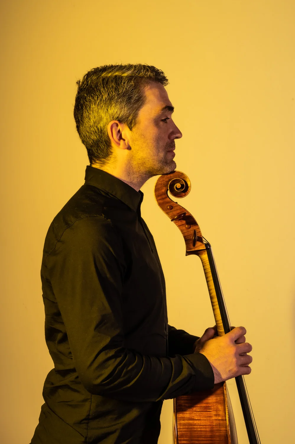 Cellist in black holding his instrument, warm yellow backdrop — Münchner Symphoniker, musician portrait photography by Janina Laszlo, Munich