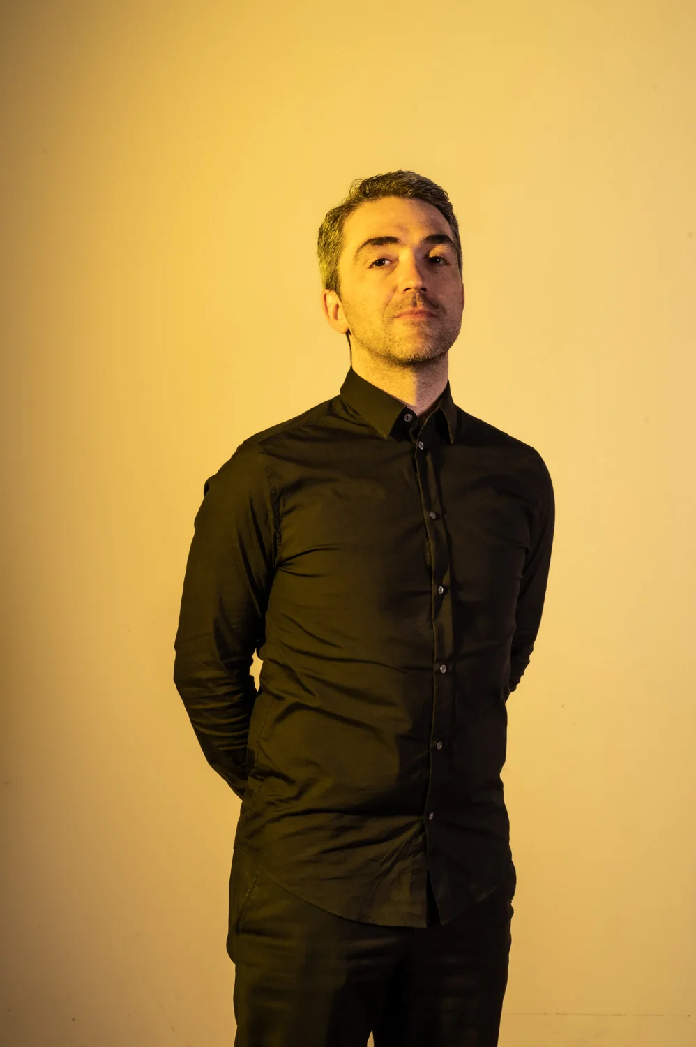 Musician in black, arms behind his back, warm yellow backdrop — Münchner Symphoniker, musician portrait photography by Janina Laszlo, Munich