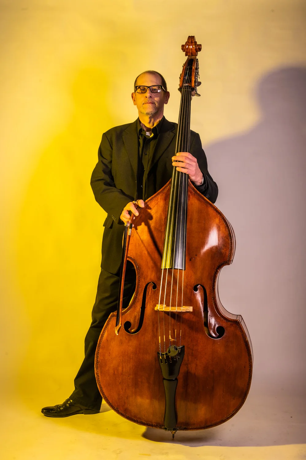 Double bassist smiling beside his instrument, warm yellow backdrop — Münchner Symphoniker, musician portrait photography by Janina Laszlo, Munich