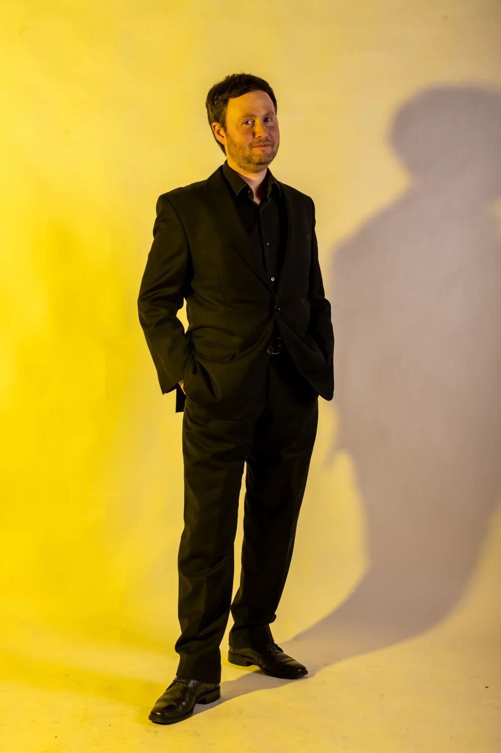 Musician in black, hands in pockets, warm yellow backdrop — Münchner Symphoniker, musician portrait photography by Janina Laszlo, Munich