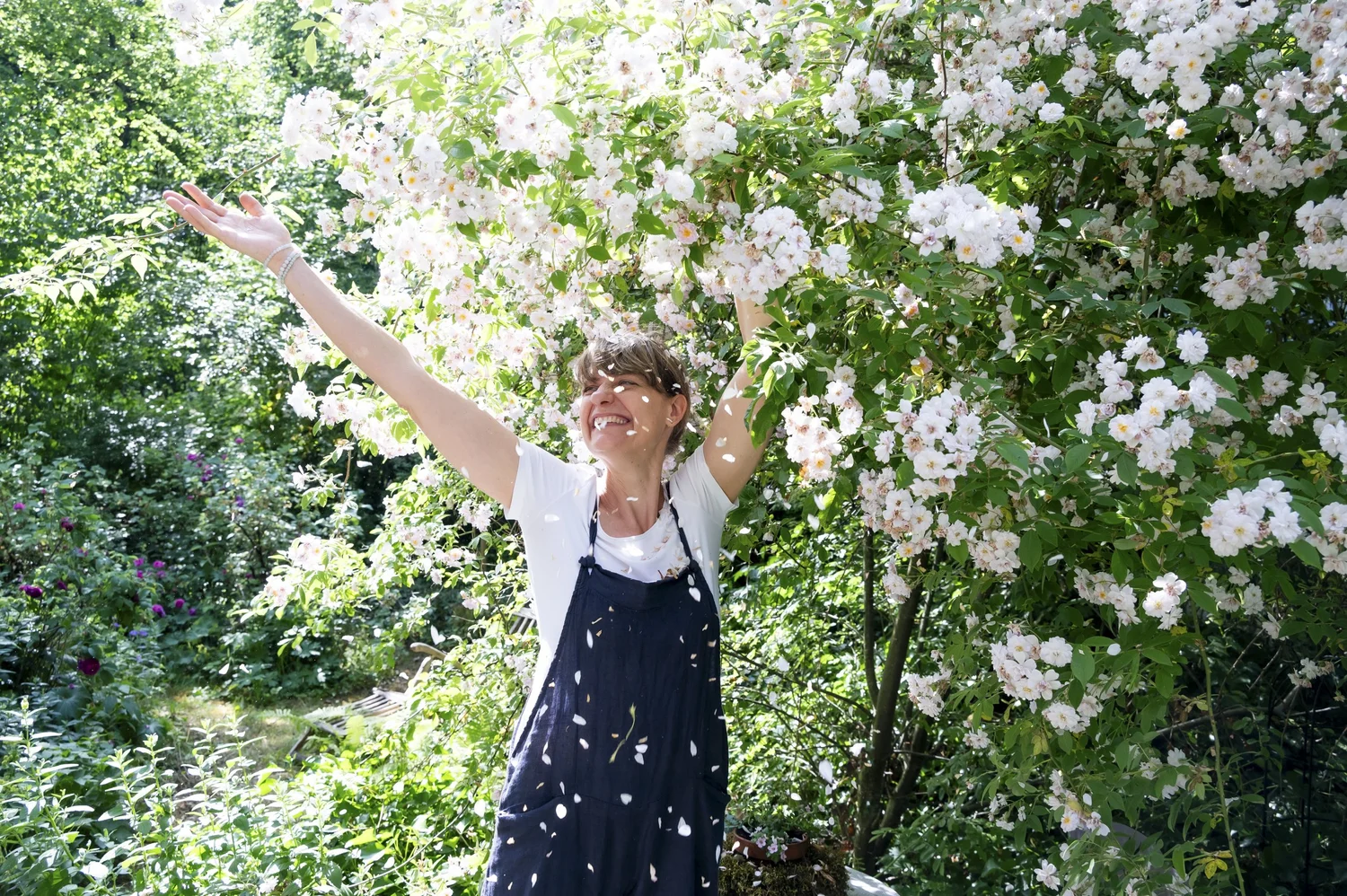 Sarah Stiller raising her arms beneath blooming white roses in her cottage garden — campaign photography by Janina Laszlo, Munich