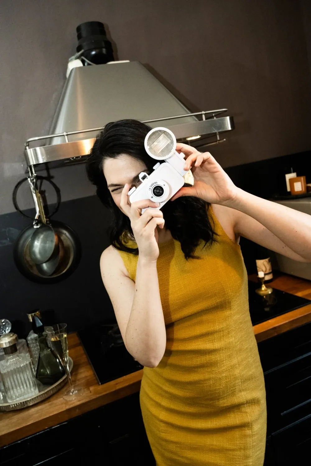 Na'ama Goldman in a yellow dress holding a vintage camera to her eye in a dark kitchen, Na'ama Goldman series — portrait by Janina Laszlo, portrait photographer in Munich