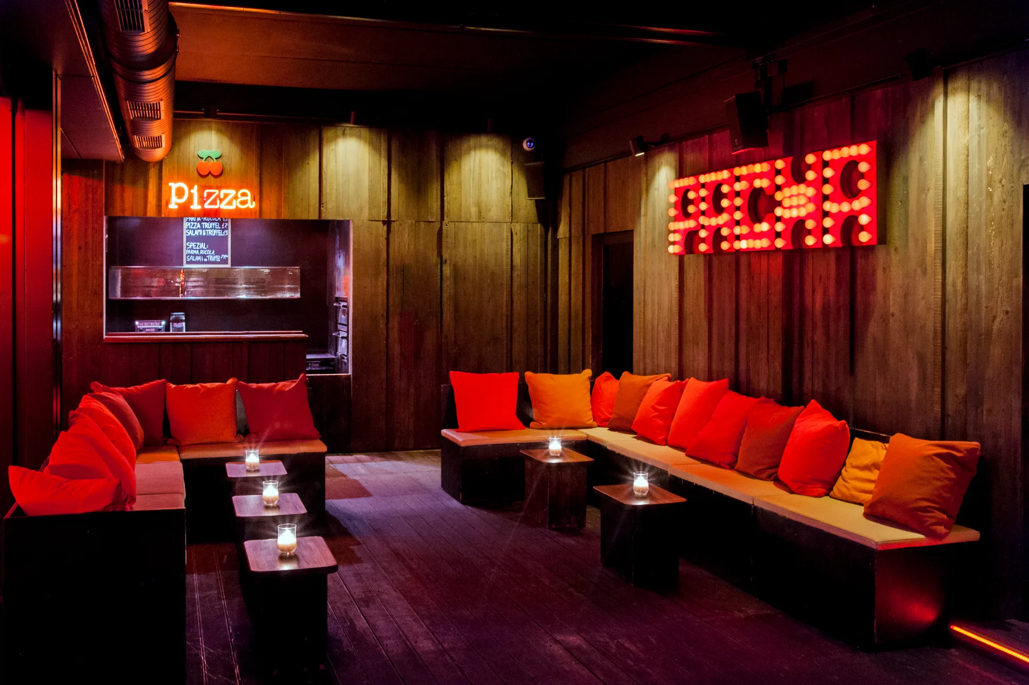 Lounge with orange and red cushions on wooden benches, neon Pizza sign and illuminated PACHA marquee letters on a dark timber wall — Pacha Munich, interior and architectural photography by Janina Laszlo, Munich