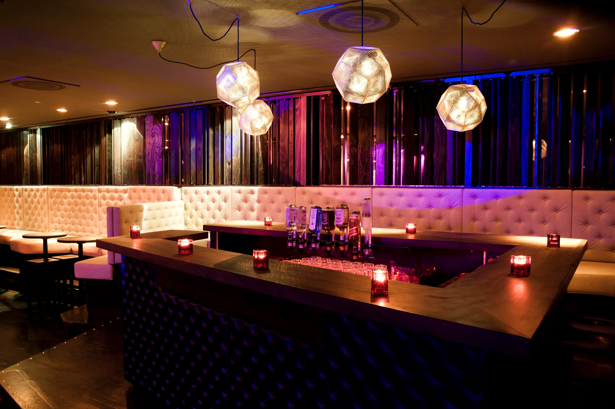 Bar with tufted white leather banquettes, brass-coloured geometric pendant lamps and violet light on dark wood panelling — Pacha Munich, interior and architectural photography by Janina Laszlo, Munich