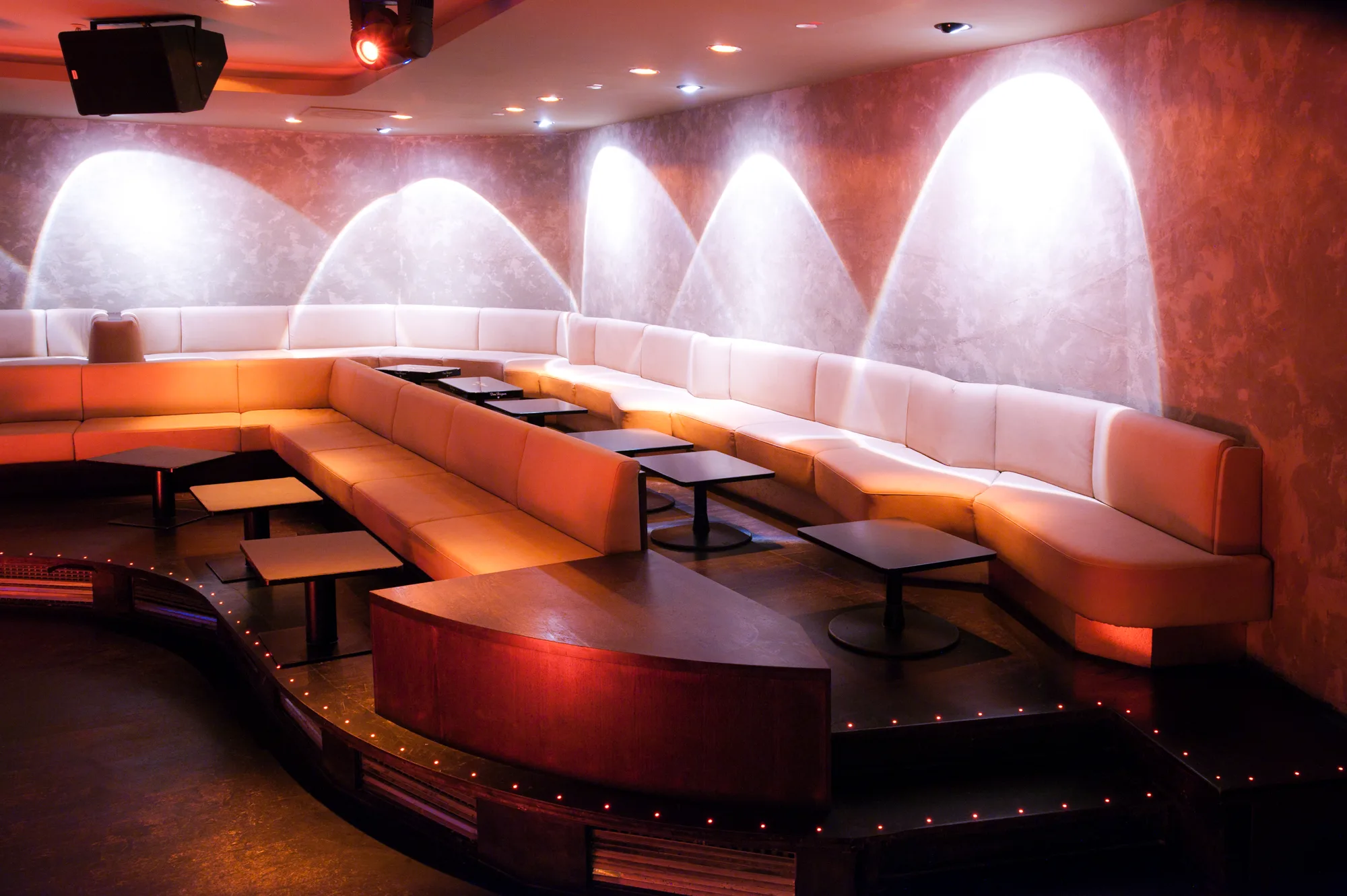 VIP lounge with curved white leather sofas, light cones on a textured plaster wall and an LED-dotted platform — Pacha Munich, interior and architectural photography by Janina Laszlo, Munich