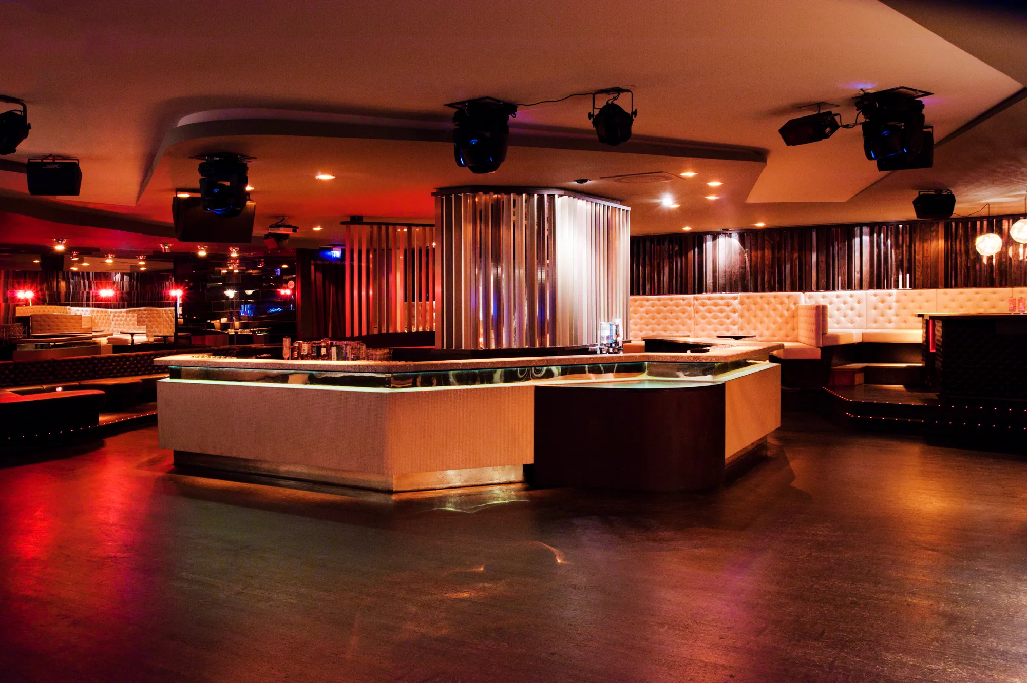 Central island bar with illuminated glass edge beneath a lamella-clad column, tufted banquettes in the background — Pacha Munich, interior and architectural photography by Janina Laszlo, Munich