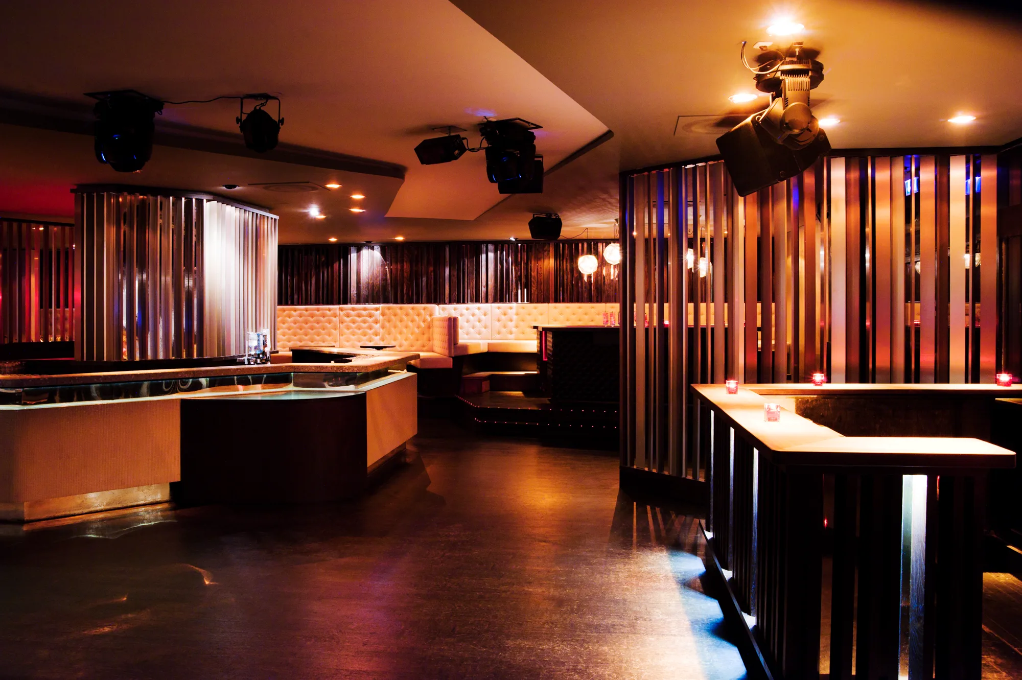Bar counter framed by bronze and wood lamella screens, tufted leather seating beyond — Pacha Munich, interior and architectural photography by Janina Laszlo, Munich