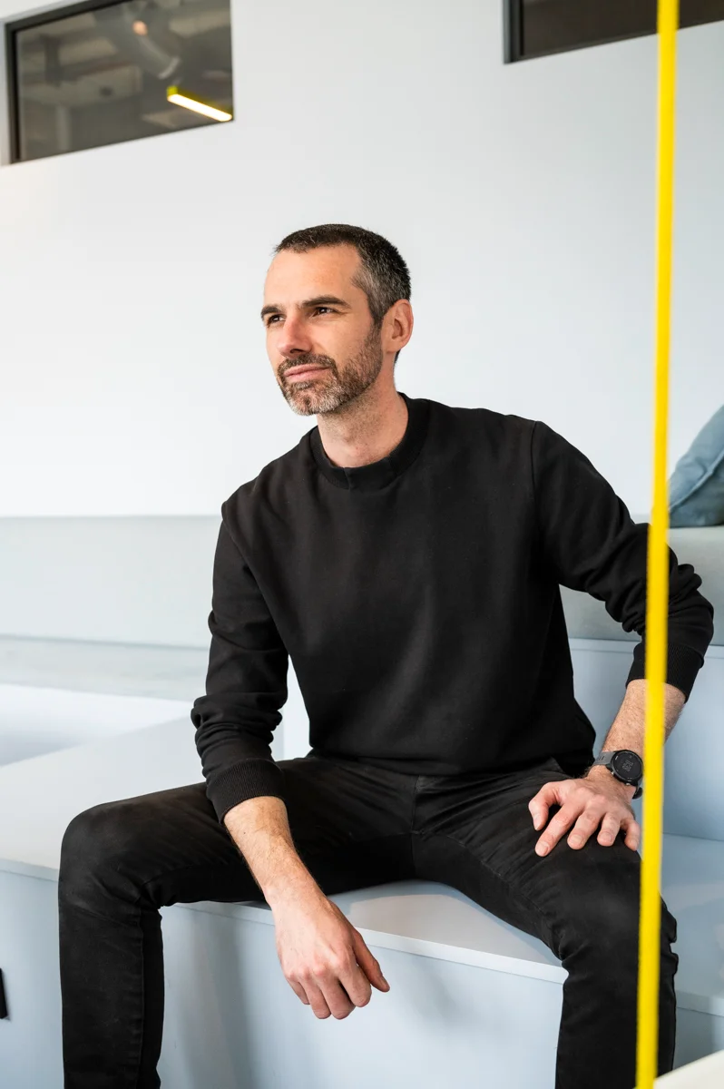 Personio co-founder in a black sweater sitting on a white bench, yellow rope in the foreground, Munich office — Personio, corporate photography by Janina Laszlo