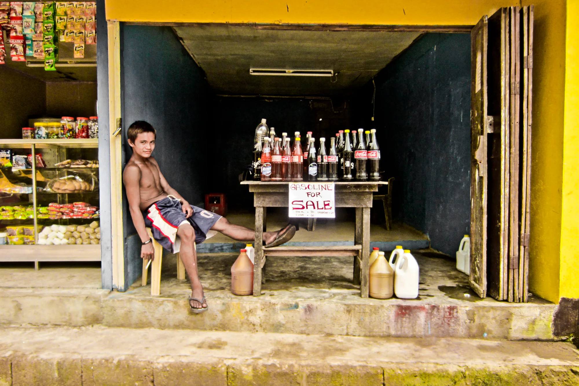 Boy leaning in a shop doorway beside bottles of gasoline for sale on a table — Palawan, Philippines, documentary and travel photography by Janina Laszlo